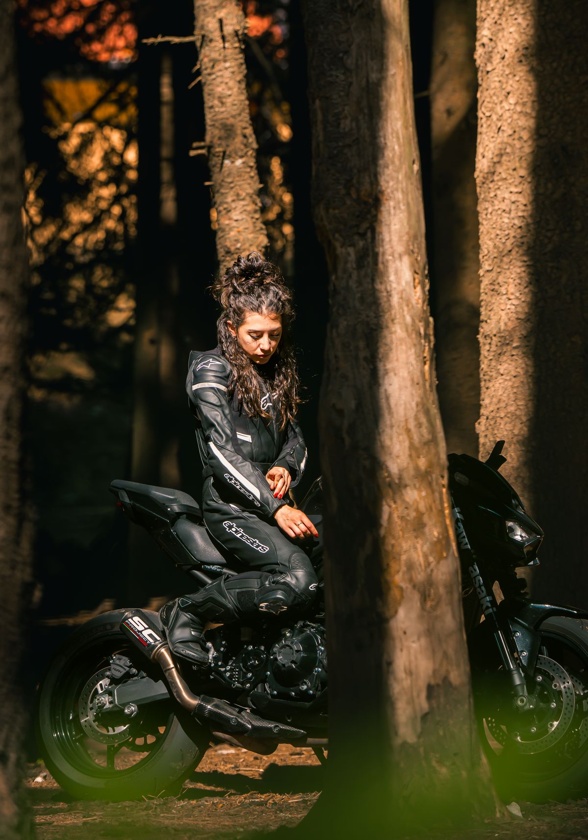 Caterina in tuta di pelle seduta sulla moto fra gli alberi